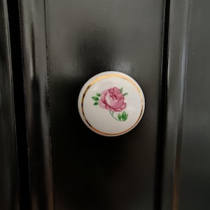 May include: A white ceramic cabinet knob with a pink rose design and gold trim. The knob is mounted on a black cabinet door. The rose has green leaves.