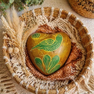 May include: A heart-shaped, hand-painted gourd with a green bird and leaf design, nestled in a woven basket. The gourd is golden brown, and the bird and leaves are painted in a vibrant green. The basket is lined with a brown and cream-colored fabric.