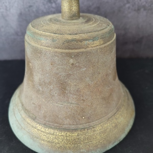 May include: A weathered brass bell with a cylindrical top handle and a flared base. The metal surface displays a dull, aged patina with visible signs of oxidation, including greenish-blue discoloration and textured, worn areas across the body.