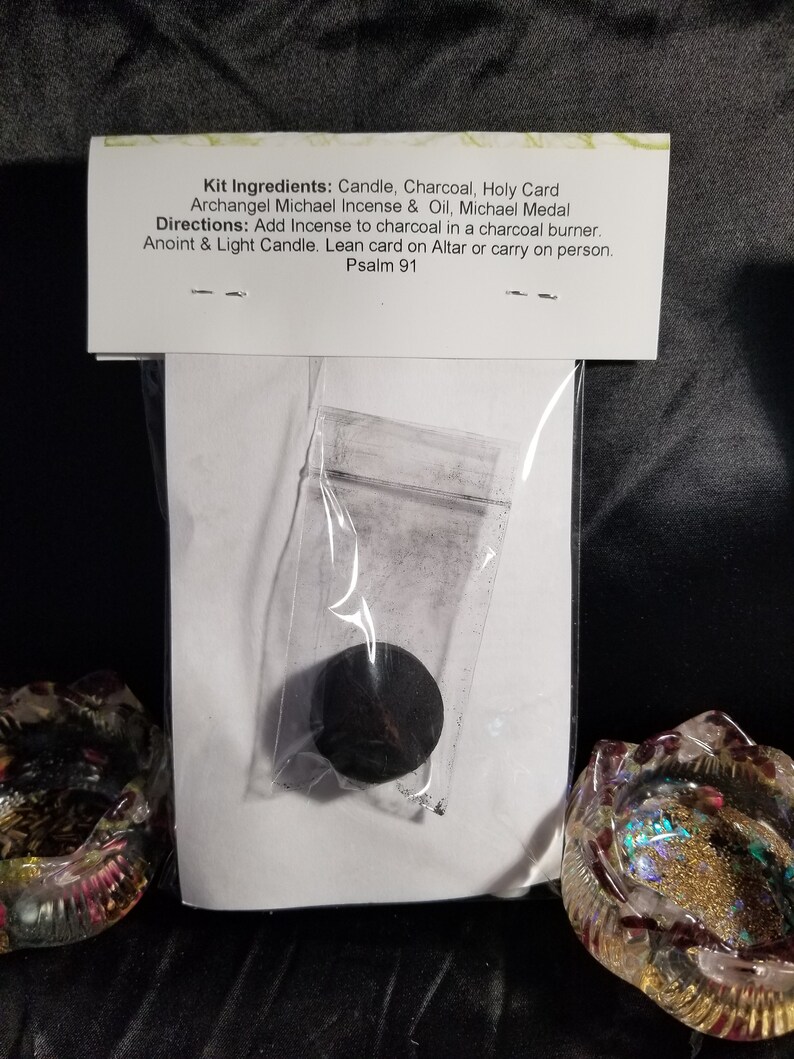 May include: A white card with the text "Kit Ingredients: Candle, Charcoal, Holy Card Archangel Michael Incense & Oil, Michael Medal Directions: Add Incense to charcoal in a charcoal burner. Anoint & Light Candle. Lean card on Altar or carry on person. Psalm 91" and a small black charcoal disc in a clear plastic bag.