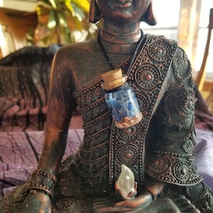 May include: A small glass bottle with a cork stopper filled with blue and gold glitter hangs from a black cord around the neck of a bronze Buddha statue.