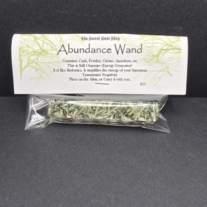 May include: A small, clear plastic bag containing a green and white crystal wand. The bag has a white label with the text "The Secret Door Shop Abundance Wand Contains: Cash, Peridot, Citrine, Amethyst, etc This is Still Orgonite (Energy Generator) It is like Radonics. It amplifies the energy of your Intention Transmutes Negativity Place on the Altar, or Carry it with you. $15"
