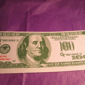 Peut inclure: Un billet de 100 dollars am&eacute;ricain vert et blanc avec un portrait de Benjamin Franklin. Le billet est marqu&eacute; "USE FOR JOSS PAPER ONLY" et porte le num&eacute;ro de s&eacute;rie "Gr10810668 D".