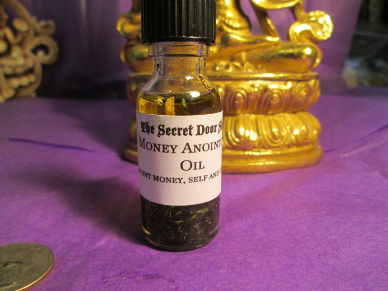 Peut inclure: Une petite bouteille en verre avec un bouchon noir remplie d'un liquide clair et d'herbes vertes. La bouteille porte une &eacute;tiquette blanche avec le texte "The Secret Door Money Anoint Oil" et "Attract Money, Self And Abundance".