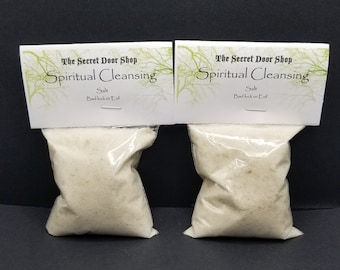 May include: Two clear plastic bags filled with white salt crystals. Each bag has a white label with the text "The Secret Door Shop" and "Spiritual Cleansing" in black text. The label also says "Salt" and "Bad luck or Evil" in black text.