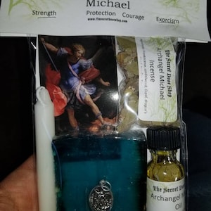 May include: A kit for Archangel Michael, featuring a painting of the angel, a white candle, a teal blue soap bar with a silver charm, a small bottle of oil with herbs, and a bag of incense. The kit is labeled "The Secret Door Shop" and includes the text "Archangel Michael" and "Protection, Courage, Exorcism".