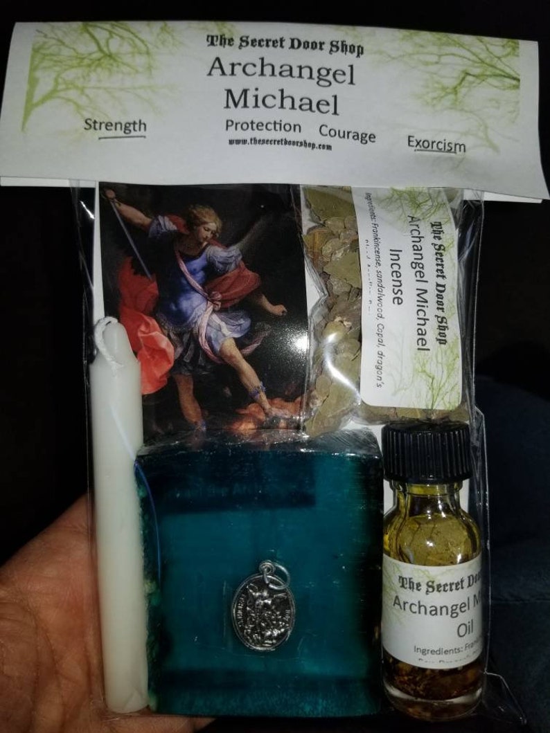 May include: A kit for Archangel Michael, featuring a painting of the angel, a white candle, a teal blue soap bar with a silver charm, a small bottle of oil with herbs, and a bag of incense. The kit is labeled "The Secret Door Shop" and includes the text "Archangel Michael" and "Protection, Courage, Exorcism".