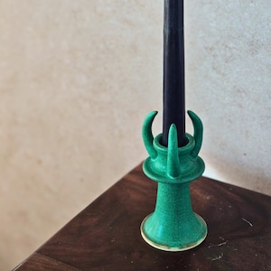 Puede incluir: Una vela negra en un candelero de cerámica verde. El candelero tiene una superficie texturizada y un diseño único con elementos curvos. La vela es alta y delgada, con una punta ligeramente curvada. El candelero está sobre una superficie de madera oscura.