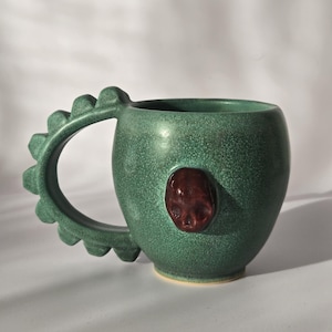 Puede incluir: Una taza de cerámica verde con una superficie texturizada y un diseño de asa único. El asa tiene una serie de puntas redondeadas. Un adorno en forma de calavera, de color rojo oscuro, está en la parte delantera. La taza está sobre un fondo blanco.