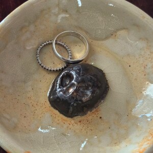 May include: A round, beige ceramic dish with a crackled glaze finish containing a dark brown, sculpted skull-shaped object and two silver-toned rings, one plain band and one thin band set with small, clear, sparkling stones.