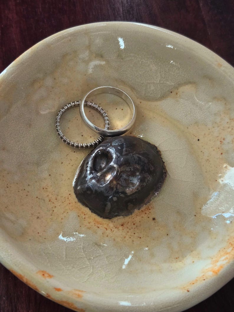 May include: A round, beige ceramic dish with a crackled glaze finish containing a dark brown, sculpted skull-shaped object and two silver-toned rings, one plain band and one thin band set with small, clear, sparkling stones.