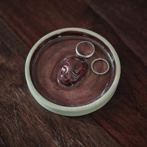 May include: A round ceramic trinket dish with a cream-colored outer rim and a deep, glossy maroon interior. A small, sculpted maroon skull is fixed to the center of the dish, accompanied by two silver-toned rings resting on the surface.