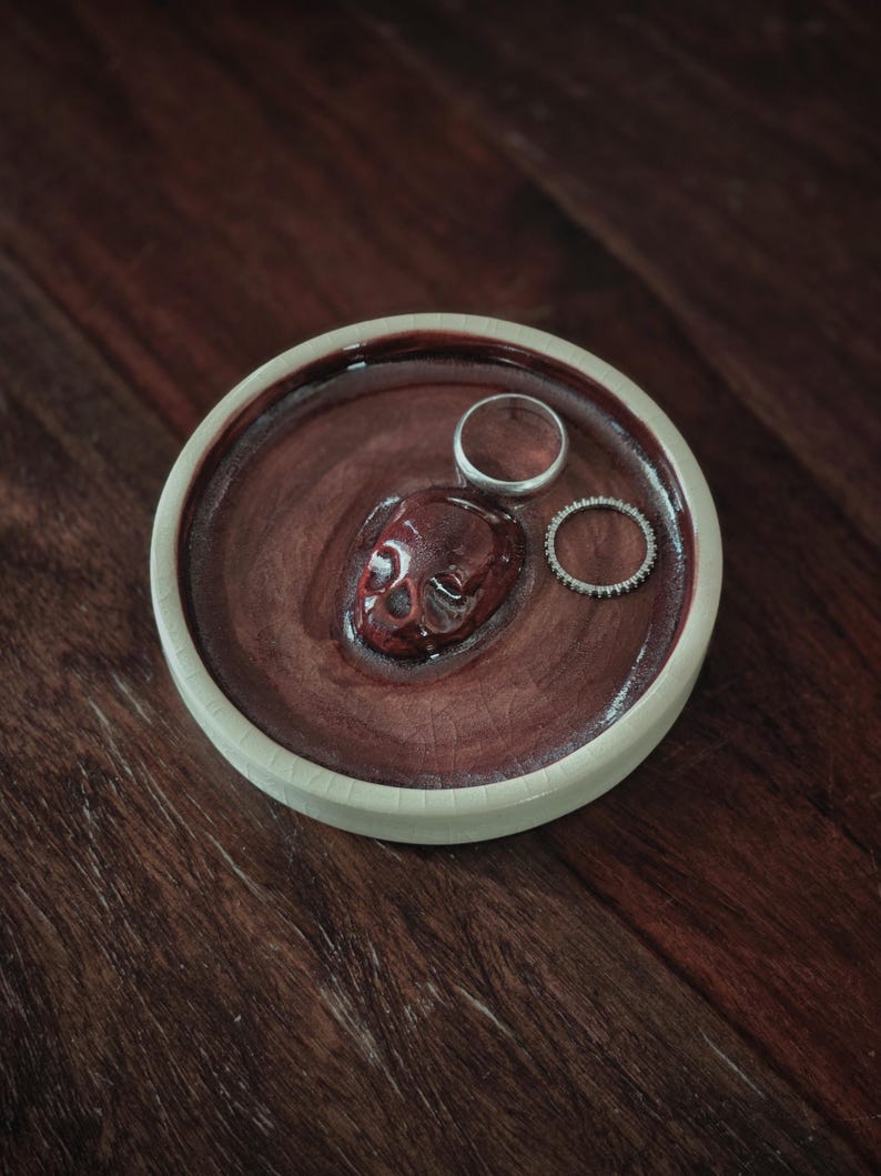 May include: A round ceramic trinket dish with a cream-colored outer rim and a deep, glossy maroon interior. A small, sculpted maroon skull is fixed to the center of the dish, accompanied by two silver-toned rings resting on the surface.