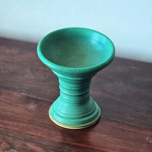K&ouml;nnte beinhalten: Eine handgefertigte, gr&uuml;ne Keramik-Sockelschale mit breitem, flachem Oberteil und geripptem, zylindrischem Sockel. Die Schale ist etwa 10 cm hoch und hat einen hellgelben Boden.