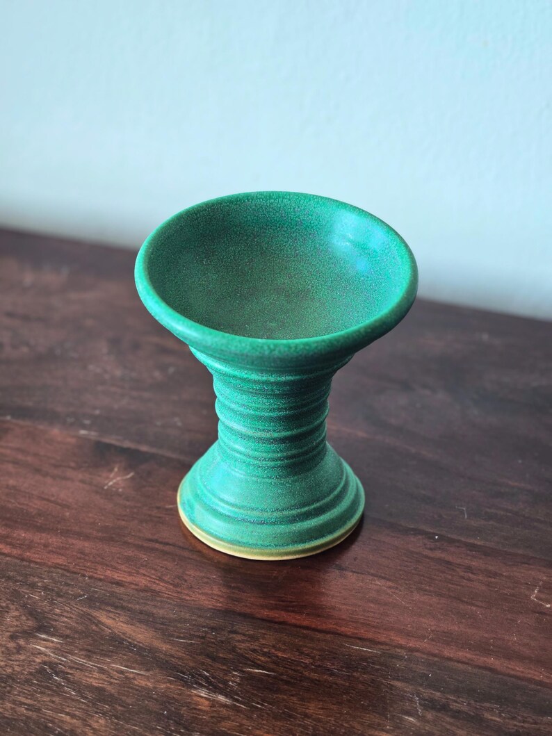 K&ouml;nnte beinhalten: Eine handgefertigte, gr&uuml;ne Keramik-Sockelschale mit breitem, flachem Oberteil und geripptem, zylindrischem Sockel. Die Schale ist etwa 10 cm hoch und hat einen hellgelben Boden.