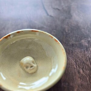 May include: A small, round, beige ceramic bowl with a glossy finish and a raised, sculpted skull figure centered at the bottom. The rim of the bowl features irregular, burnt-orange accents, and the interior surface has a smooth, glazed texture.