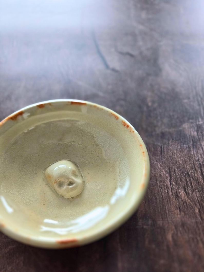 May include: A small, round, beige ceramic bowl with a glossy finish and a raised, sculpted skull figure centered at the bottom. The rim of the bowl features irregular, burnt-orange accents, and the interior surface has a smooth, glazed texture.