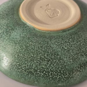 May include: A ceramic bowl with a mottled green glaze featuring a textured, speckled surface. The underside reveals a smooth, unglazed beige circular base with a stamped maker's mark depicting a small teapot icon with the letters XP inside.