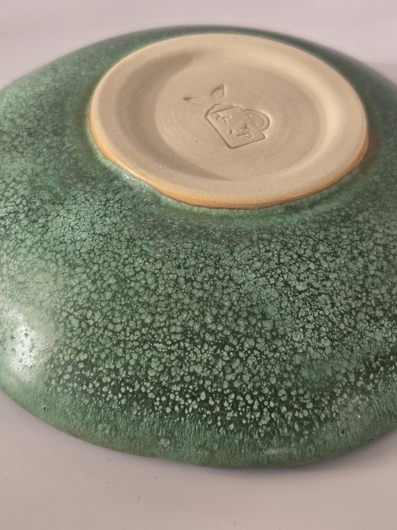 May include: A ceramic bowl with a mottled green glaze featuring a textured, speckled surface. The underside reveals a smooth, unglazed beige circular base with a stamped maker's mark depicting a small teapot icon with the letters XP inside.