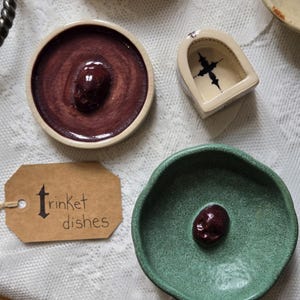 May include: A collection of ceramic trinket dishes including a round dish with a dark red crackle glaze and a central skull, a green textured dish with a dark red skull, and a small arched dish with a black cross. A brown tag reads trinket dishes.