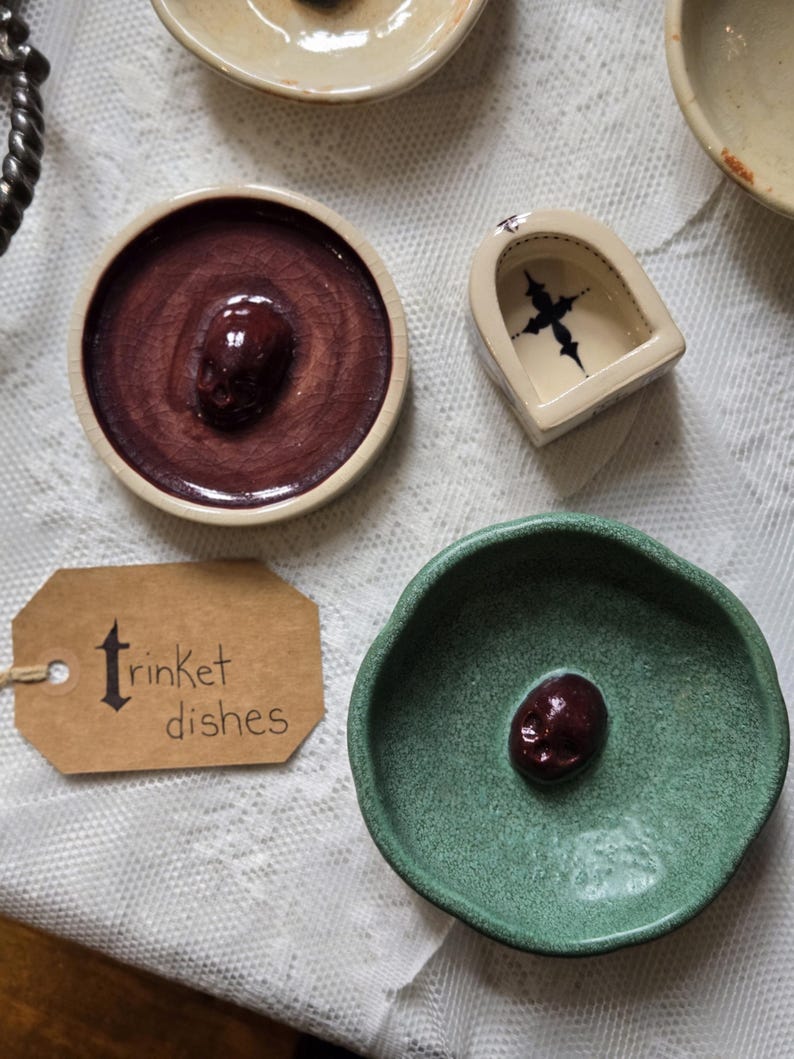 May include: A collection of ceramic trinket dishes including a round dish with a dark red crackle glaze and a central skull, a green textured dish with a dark red skull, and a small arched dish with a black cross. A brown tag reads trinket dishes.