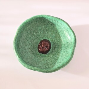 May include: A small, round, handmade ceramic bowl with a vibrant, textured green glaze. The interior of the bowl features a single, dark brown, glossy skull figurine embedded in the center, creating a stark contrast against the bright green surface.