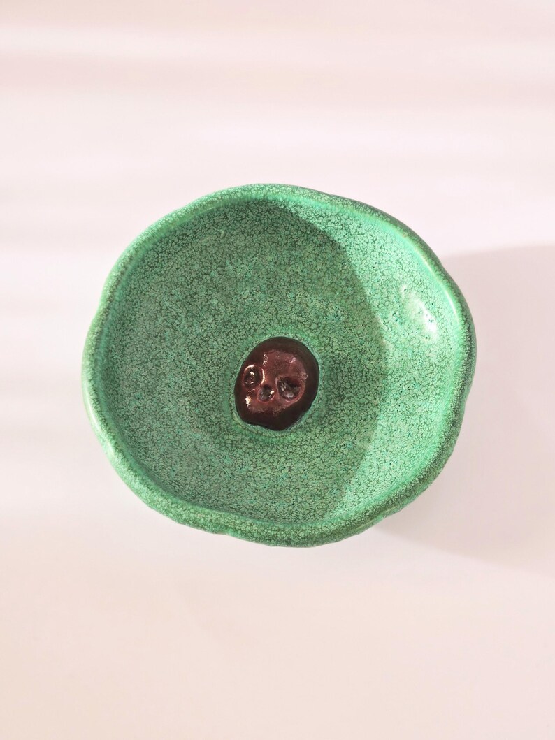 May include: A small, round, handmade ceramic bowl with a vibrant, textured green glaze. The interior of the bowl features a single, dark brown, glossy skull figurine embedded in the center, creating a stark contrast against the bright green surface.