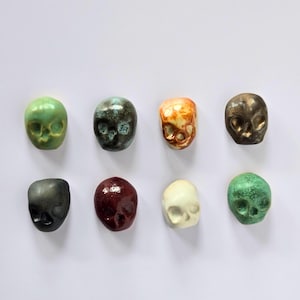 May include: Eight small ceramic skull figurines in a variety of colours. The skulls are arranged in two rows of four, with colours including green, black, brown, and white. Each skull has detailed facial features.
