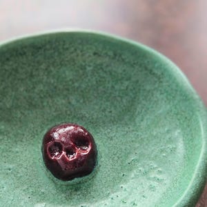 May include: A small, round, green ceramic bowl with a textured, crackled glaze finish. Centered at the bottom of the bowl is a small, dark maroon, glossy skull-shaped object with visible eye sockets and a nasal cavity, resting on the surface.