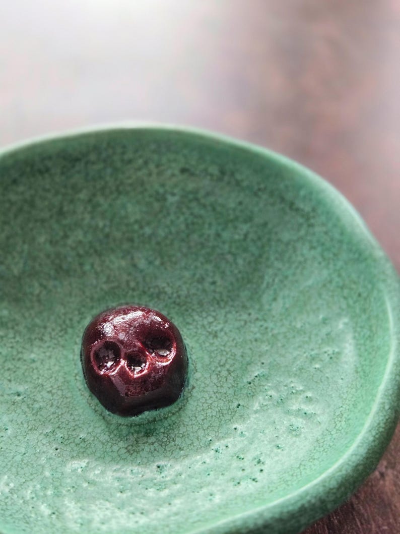 May include: A small, round, green ceramic bowl with a textured, crackled glaze finish. Centered at the bottom of the bowl is a small, dark maroon, glossy skull-shaped object with visible eye sockets and a nasal cavity, resting on the surface.