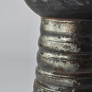 K&ouml;nnte beinhalten: Nahaufnahme einer Keramikvase mit strukturierter, metallischer Oberfl&auml;che. Die Vase hat einen bauchigen oberen Teil und einen geriffelten, zylindrischen Sockel. Die Farbpalette umfasst Grau-, Silber- und Bronzet&ouml;ne.