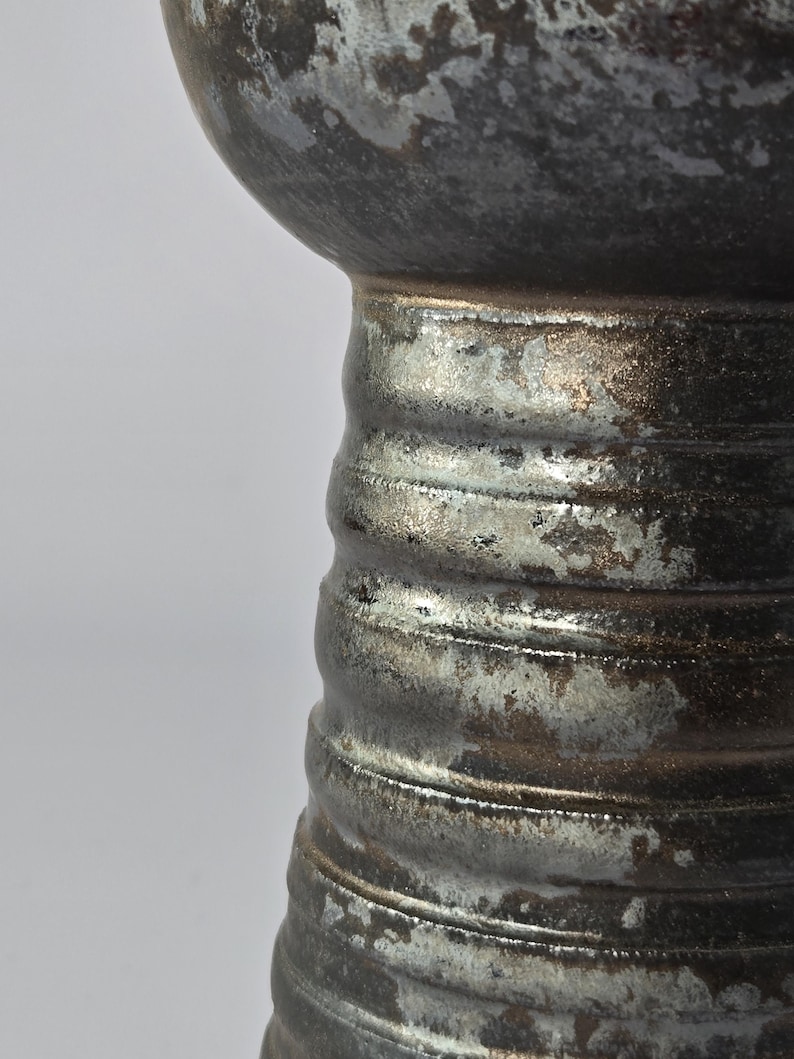 K&ouml;nnte beinhalten: Nahaufnahme einer Keramikvase mit strukturierter, metallischer Oberfl&auml;che. Die Vase hat einen bauchigen oberen Teil und einen geriffelten, zylindrischen Sockel. Die Farbpalette umfasst Grau-, Silber- und Bronzet&ouml;ne.