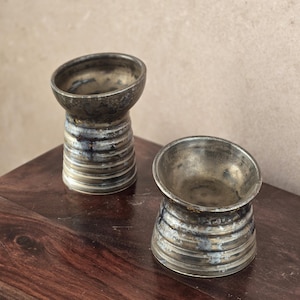 K&ouml;nnte beinhalten: Zwei handgefertigte Keramikgef&auml;&szlig;e in Metallic-T&ouml;nen mit einem einzigartigen, abgewinkelten Rand. Die Gef&auml;&szlig;e haben einen zylindrischen Sockel mit horizontalen Rillen und eine dunkle, gesprenkelte Oberfl&auml;che. Sie stehen auf einer dunklen Holzoberfl&auml;che und zeigen ihre handwerkliche Qualit&auml;t.