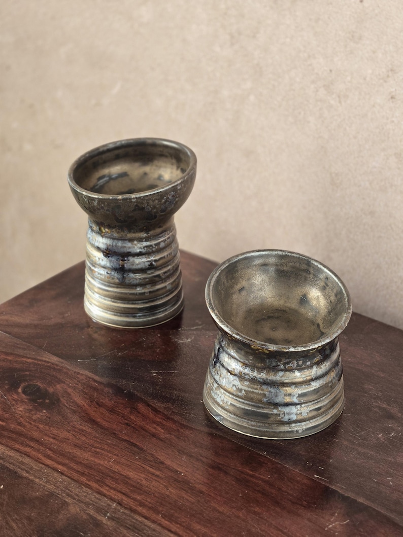 K&ouml;nnte beinhalten: Zwei handgefertigte Keramikgef&auml;&szlig;e in Metallic-T&ouml;nen mit einem einzigartigen, abgewinkelten Rand. Die Gef&auml;&szlig;e haben einen zylindrischen Sockel mit horizontalen Rillen und eine dunkle, gesprenkelte Oberfl&auml;che. Sie stehen auf einer dunklen Holzoberfl&auml;che und zeigen ihre handwerkliche Qualit&auml;t.