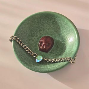 May include: A round, green ceramic dish with a crackled glaze finish featuring a small, dark red, three-dimensional skull at its center. A silver-toned metal chain bracelet with a single iridescent, teardrop-shaped crystal charm rests inside.