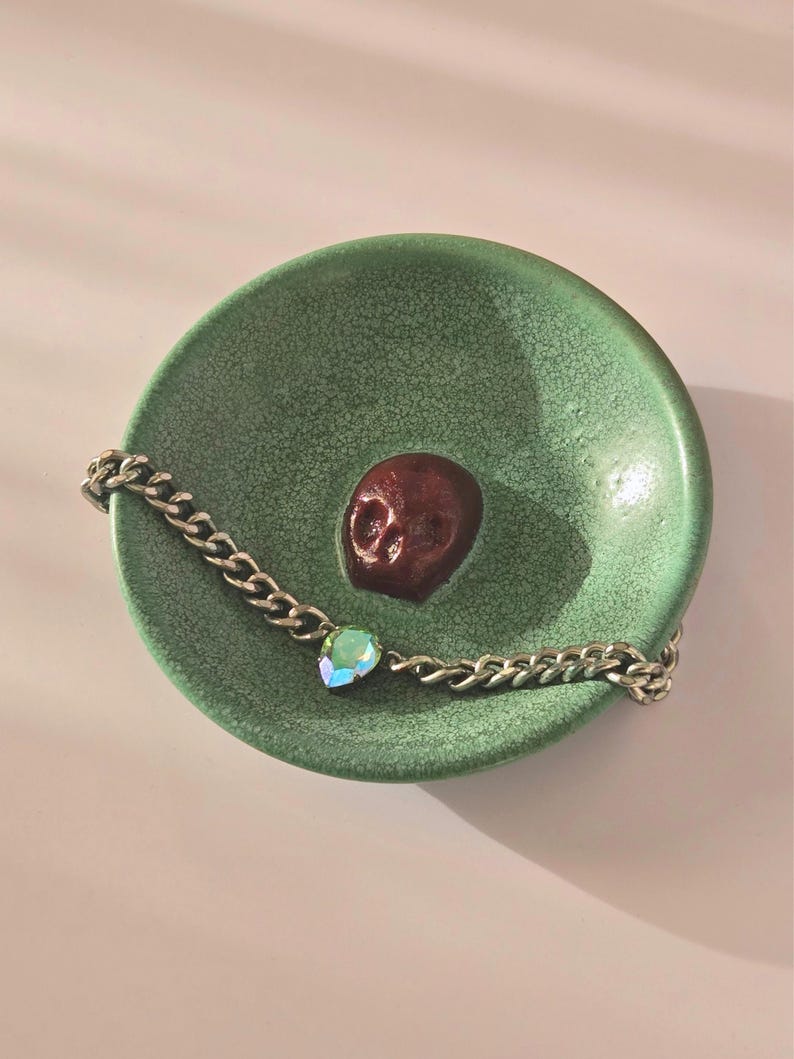 May include: A round, green ceramic dish with a crackled glaze finish featuring a small, dark red, three-dimensional skull at its center. A silver-toned metal chain bracelet with a single iridescent, teardrop-shaped crystal charm rests inside.