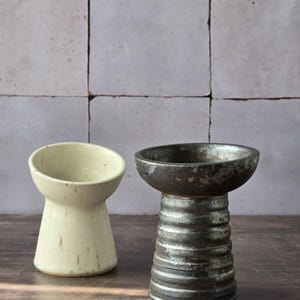 K&ouml;nnte beinhalten: Zwei Keramikgef&auml;&szlig;e mit minimalistischem Design. Eines ist cremefarben mit ausgestellter Oberseite, das andere ist dunkelgrau mit gerippter Basis. Beide stehen auf einer Holzoberfl&auml;che vor einem grauen Fliesen-Hintergrund.
