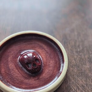May include: A circular ceramic dish featuring a deep burgundy, crackled glaze finish. The center of the dish is adorned with a raised, three-dimensional skull sculpture in a matching glossy dark red tone, surrounded by a smooth, light cream-colored rim.