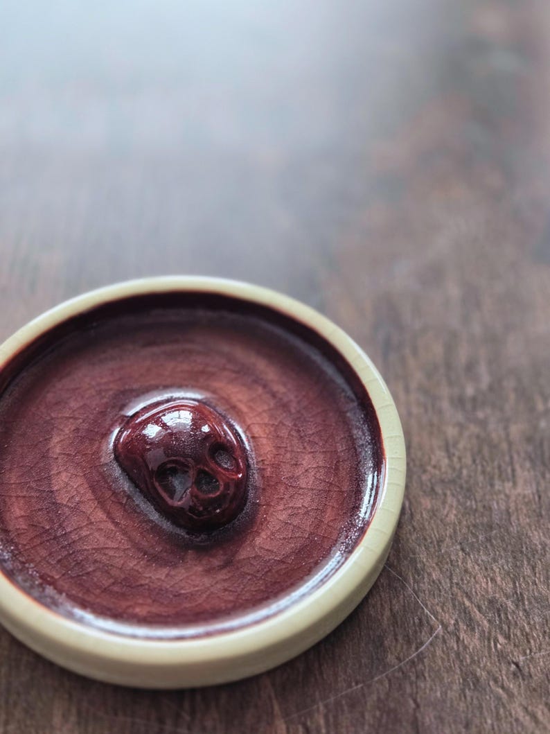 May include: A circular ceramic dish featuring a deep burgundy, crackled glaze finish. The center of the dish is adorned with a raised, three-dimensional skull sculpture in a matching glossy dark red tone, surrounded by a smooth, light cream-colored rim.