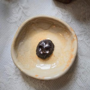 May include: A small, round, off-white ceramic bowl with a glossy, crackled glaze finish. Centered at the bottom of the bowl is a dark, metallic-toned, sculpted skull ornament with visible eye sockets and a nose cavity, resting on the interior surface.