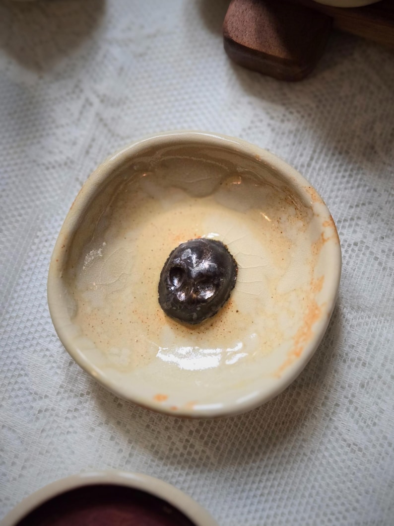 May include: A small, round, off-white ceramic bowl with a glossy, crackled glaze finish. Centered at the bottom of the bowl is a dark, metallic-toned, sculpted skull ornament with visible eye sockets and a nose cavity, resting on the interior surface.
