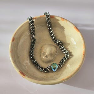 May include: A silver-toned metal chain necklace featuring a single pear-shaped iridescent crystal pendant. The necklace is resting inside a small, round, beige ceramic bowl that has a molded skull relief at the center of its interior base.