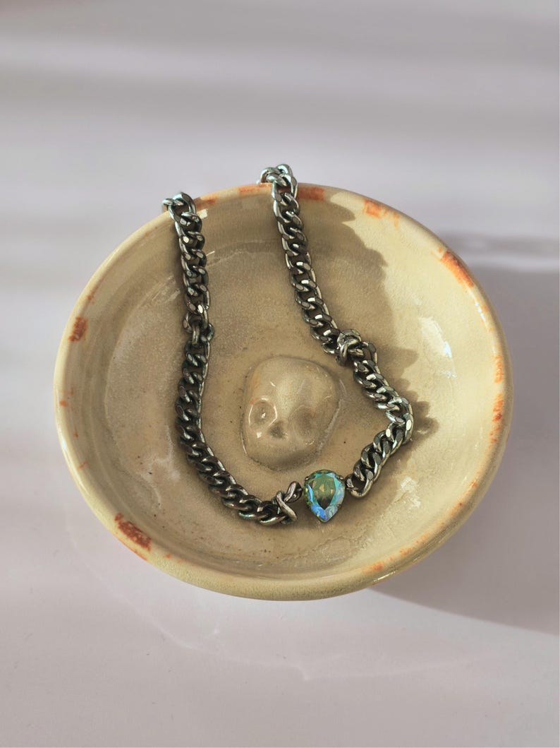 May include: A silver-toned metal chain necklace featuring a single pear-shaped iridescent crystal pendant. The necklace is resting inside a small, round, beige ceramic bowl that has a molded skull relief at the center of its interior base.