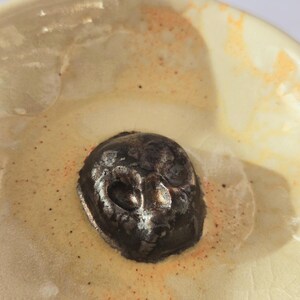 May include: A ceramic bowl with a glossy cream-colored glaze featuring orange speckles and a dark, metallic, skull-shaped sculpture fixed in the center. The bowl has a smooth, rounded interior surface with visible fine crackle patterns in the glaze.