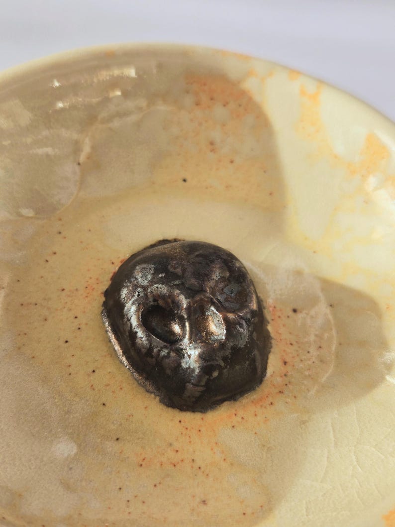May include: A ceramic bowl with a glossy cream-colored glaze featuring orange speckles and a dark, metallic, skull-shaped sculpture fixed in the center. The bowl has a smooth, rounded interior surface with visible fine crackle patterns in the glaze.