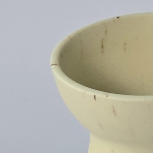 K&ouml;nnte beinhalten: Nahaufnahme einer cremefarbenen Keramikvase mit ausgestelltem Rand. Die Vase hat einen abgerundeten K&ouml;rper und einen schmalen Boden. Auf der Oberfl&auml;che befinden sich kleine braune Flecken. Der Hintergrund ist einfarbig wei&szlig;.