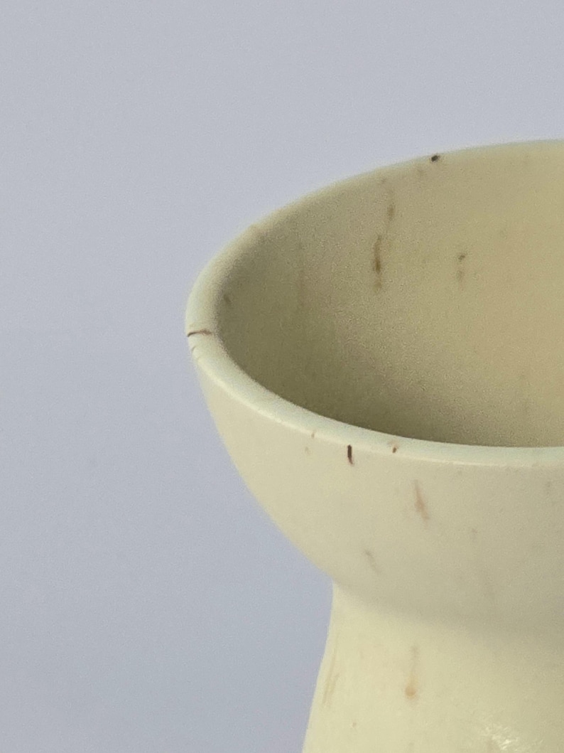 K&ouml;nnte beinhalten: Nahaufnahme einer cremefarbenen Keramikvase mit ausgestelltem Rand. Die Vase hat einen abgerundeten K&ouml;rper und einen schmalen Boden. Auf der Oberfl&auml;che befinden sich kleine braune Flecken. Der Hintergrund ist einfarbig wei&szlig;.