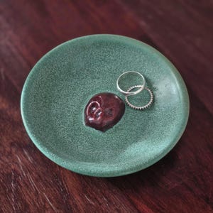 May include: A round, sage green ceramic trinket dish with a crackled glaze finish. A small, dark red, sculpted skull is attached to the center of the dish. Two thin, silver-colored metal rings rest on the surface of the dish next to the skull.