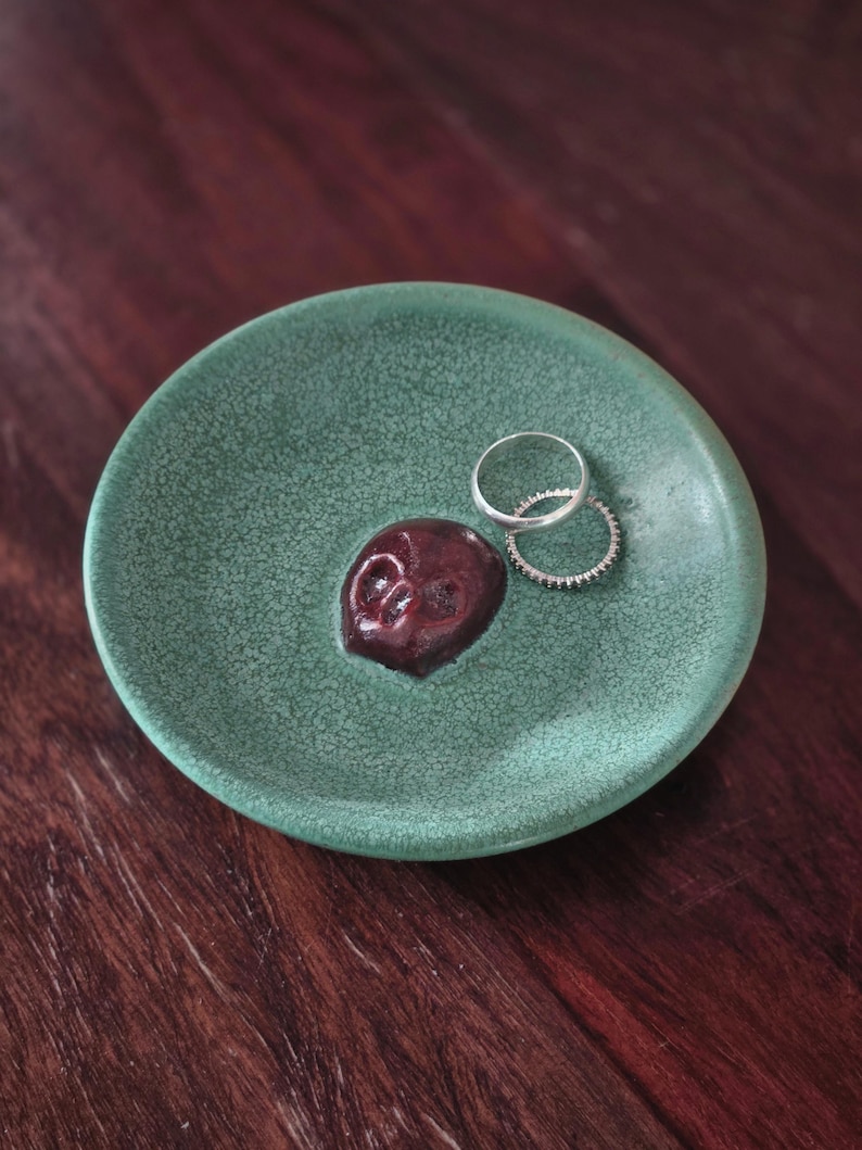 May include: A round, sage green ceramic trinket dish with a crackled glaze finish. A small, dark red, sculpted skull is attached to the center of the dish. Two thin, silver-colored metal rings rest on the surface of the dish next to the skull.