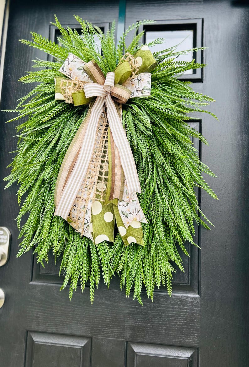 May include: A green wreath with a brown and white striped ribbon, a green polka dot ribbon, and a brown and green burlap ribbon. The wreath is hanging on a black door.